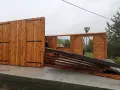 Drewniana wiata garażowa bez pozwolenia w trakcie budowy. Widać fragmenty ścian i bramy, a także stos desek.