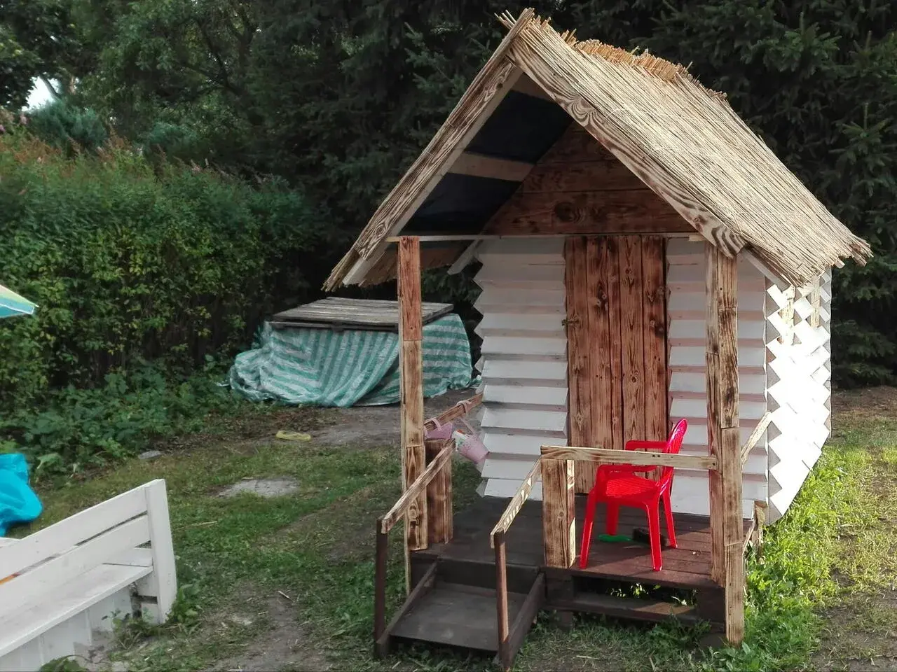 Domek dla dzieci z palet, z drewnianymi drzwiami i słomianym dachem, gotowy na zabawę.