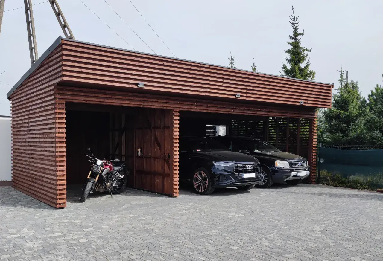Drewniana wiata na samochód przed domem, chroniąca dwa czarne SUV-y i motocykl.