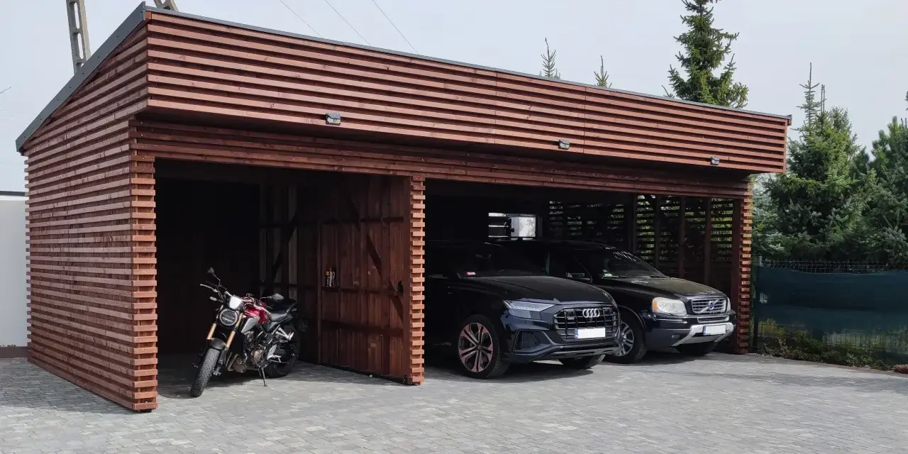 Drewniana wiata na samochód przed domem, chroniąca dwa czarne SUV-y i motocykl.