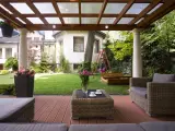 Drewniana pergola z przezroczystym dachem, tworząca zacienione miejsce do wypoczynku na tarasie. W tle ogród z huśtawką i piaskownicą.