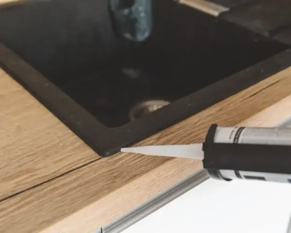 Pistolet z silikonem aplikuje uszczelnienie wokół zlewu, pokazując, jak polożyć silikon na blacie kuchennym.