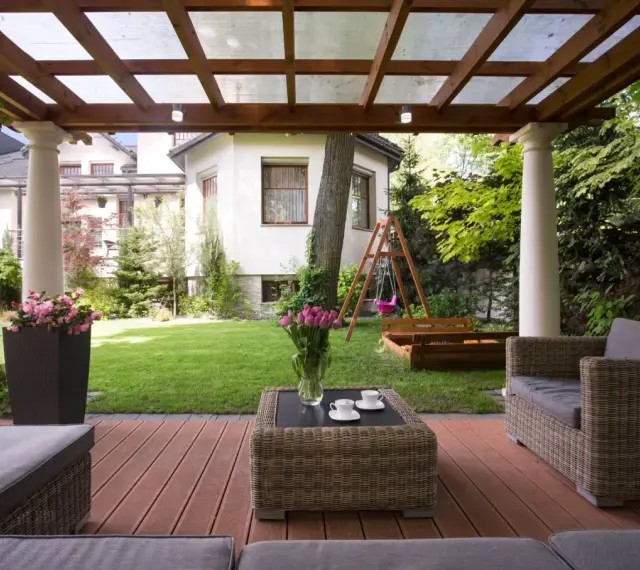 Drewniana pergola z przezroczystym dachem, tworząca zacienione miejsce do wypoczynku na tarasie. W tle ogród z huśtawką i piaskownicą.