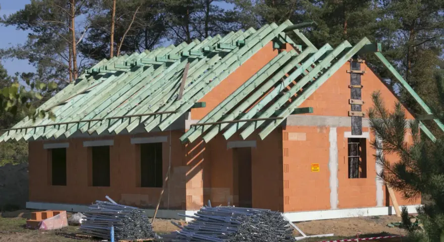 Budowa domu w metodzie gospodarczej: wzniesione ściany z pustaków ceramicznych, gotowy szkielet dachu z zielonych krokwi, materiały budowlane na placu.