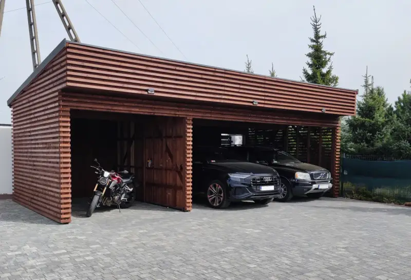 Drewniana wiata na samochód przed domem, chroniąca dwa czarne SUV-y i motocykl.
